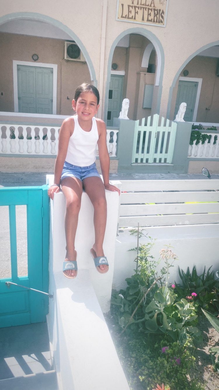 A child sitting on a whitewashed wall outside a Greek villa with mint green shutters