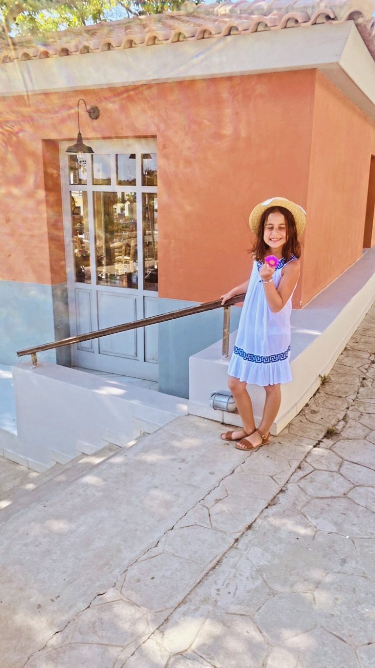 A child in a blue and white dress and straw hat outside a terracotta taverna in Greece