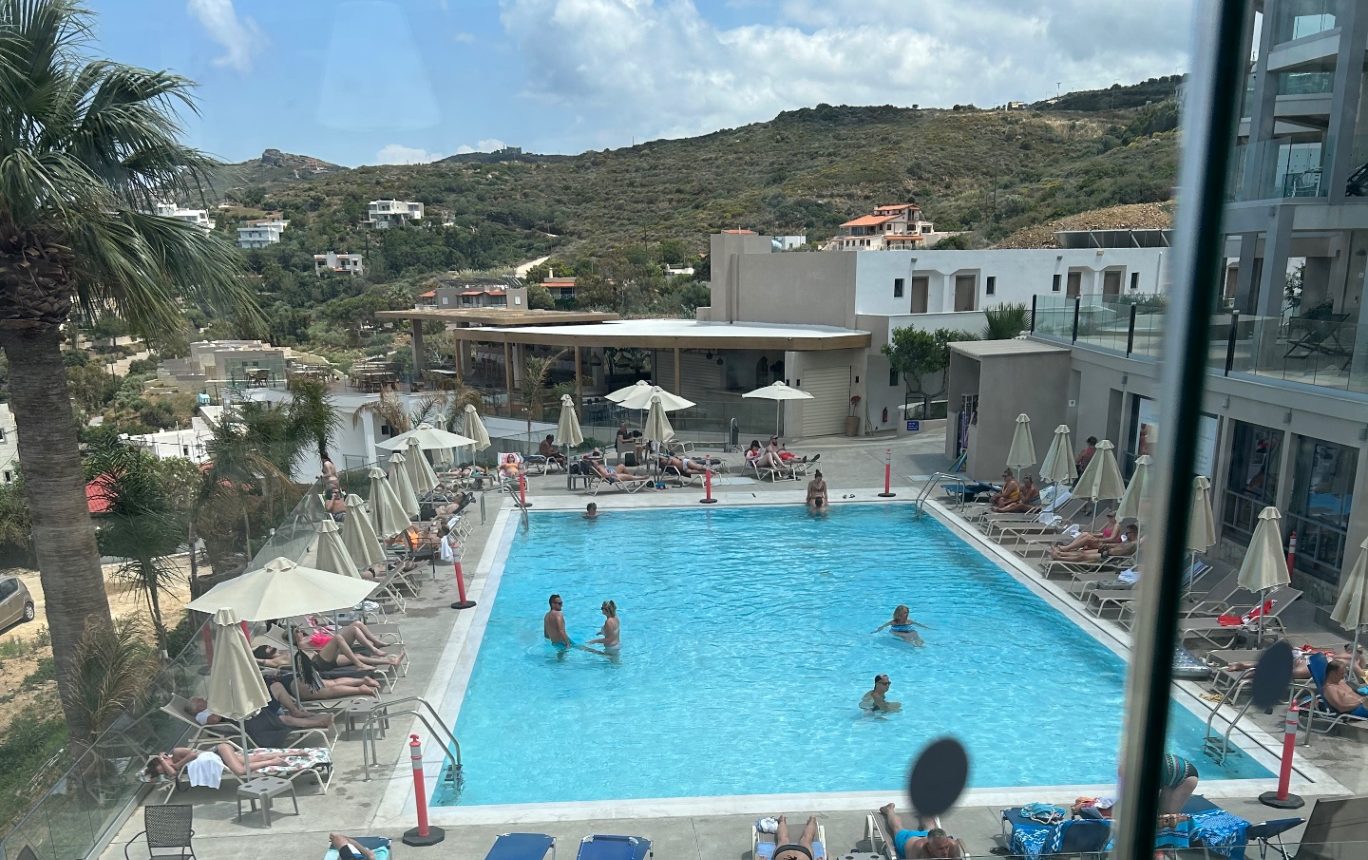 A bright blue Greek hillside hotel pool surrounded by sun lounges, umbrellas and palm trees