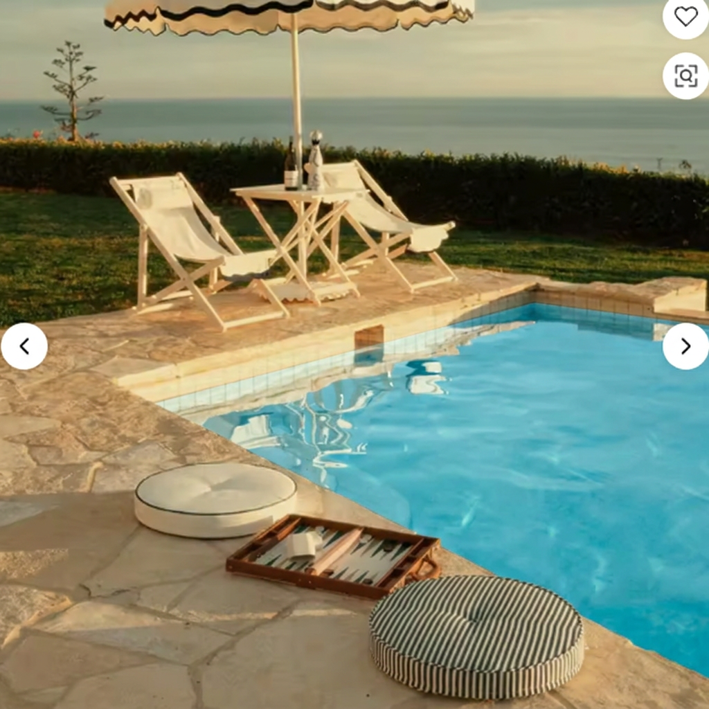 Round striped and white poolside cushions beside a stone pool at golden hour, with deck chairs and a striped umbrella